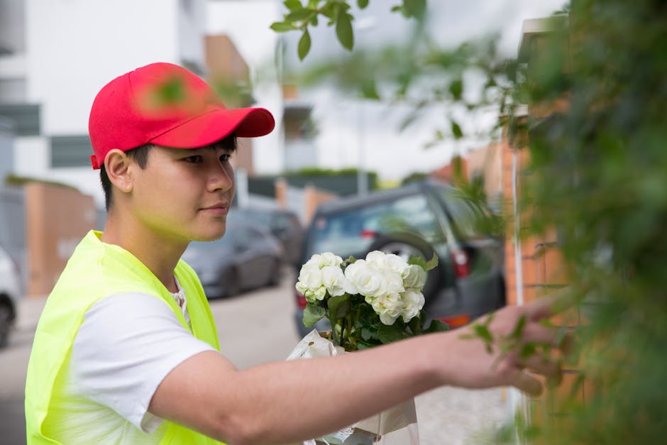 A delivery person wearing a red cap and a neon yellow high-visibility vest is holding a bouquet of fresh white roses and green foliage. The flowers are carefully arranged and wrapped in light-colored