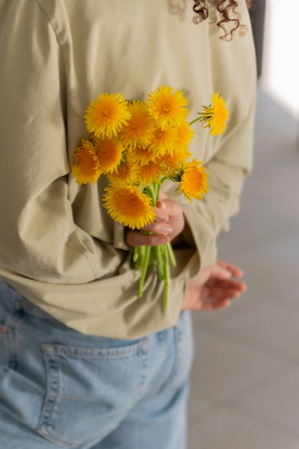 A person holding a bouquet of vibrant yellow daisies with long green stems, wrapped in clear plastic, against a soft, neutral background. The individual is wearing a beige jacket and light blue jeans,