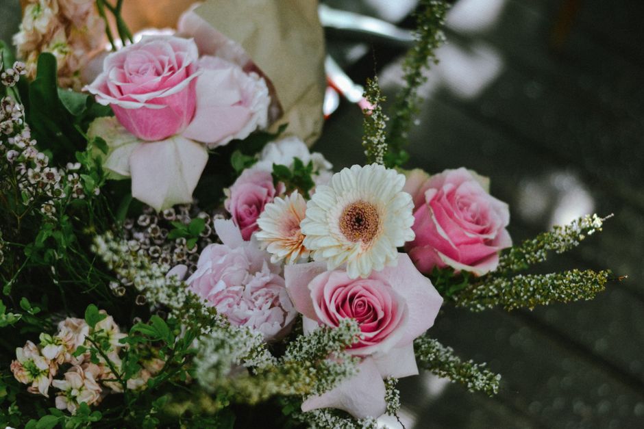 A close-up of a floral arrangement featuring soft pink roses, white gerbera daisies with dark centers, and delicate white and pink filler flowers. The bouquet is lush and vibrant, with fresh, velvety