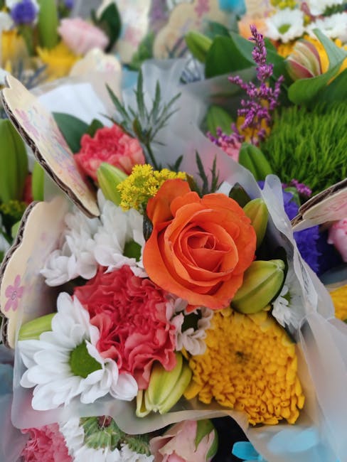 A vibrant floral arrangement featuring an orange rose at the center, surrounded by white daisies with green centers, pink carnations, yellow chrysanthemum, and purple statice. The flowers are wrapped