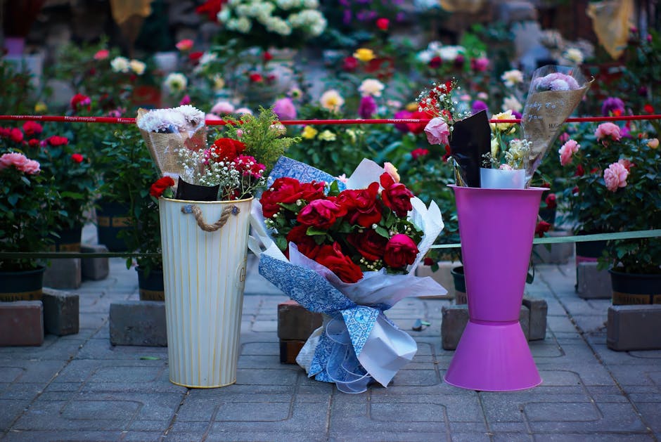A close-up of three floral arrangements placed outdoors on a paved surface in front of a background filled with blooming pink, red, and white roses. The center bouquet features deep red roses wrapped