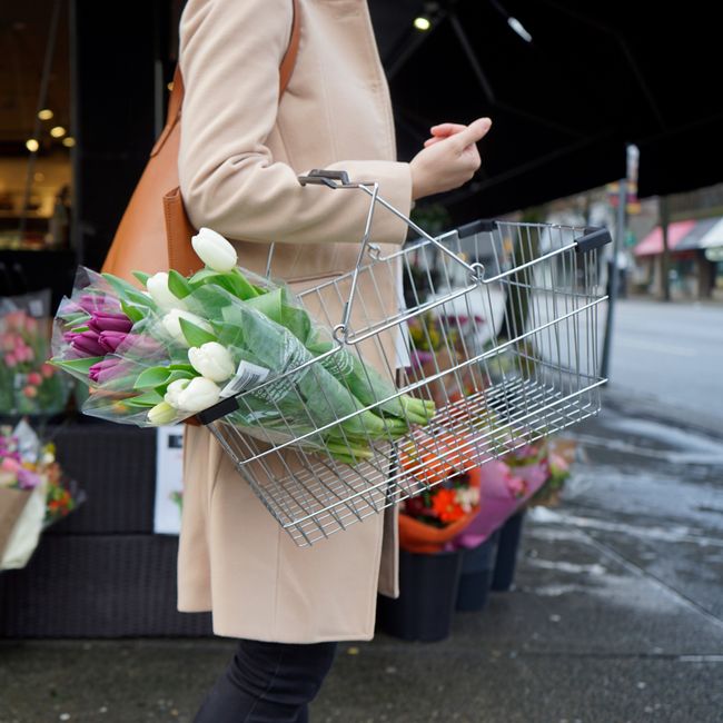 A person wearing a beige coat is holding a metal shopping basket containing a bouquet of fresh tulips in white and purple, wrapped with clear plastic. The bouquet is positioned at the side of the bask