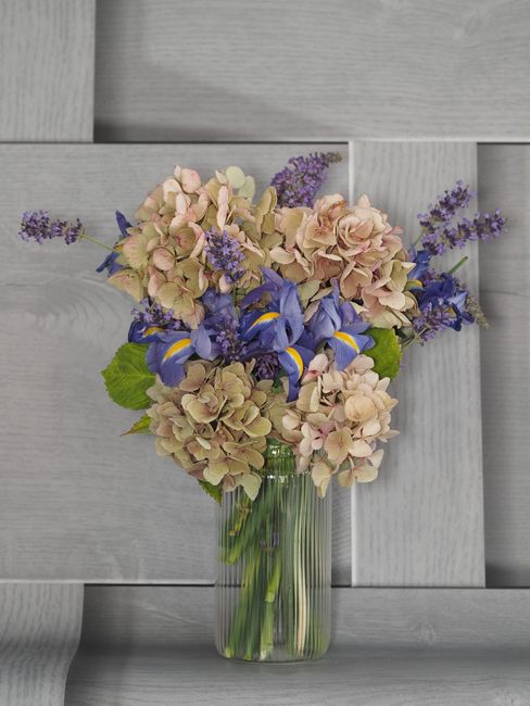 A floral arrangement featuring a bouquet of pastel pink and cream hydrangeas, vibrant purple irises, and sprigs of delicate lavender. The flowers are displayed in a clear glass vase, with green stems 
